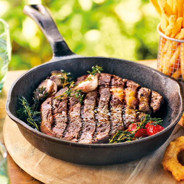 Grilled steak with vegetables in a cast iron skillet on a wooden surface with a blurred green background