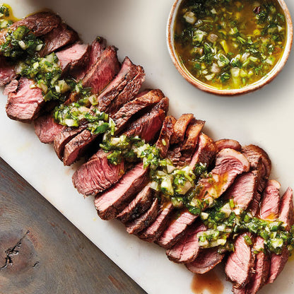 Sliced steak with green herb sauce on a white plate with a bowl of sauce in the background.