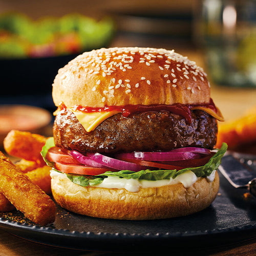 Hamburger with cheese, lettuce, tomato, and onion on a sesame seed bun with fries and salad in the background.